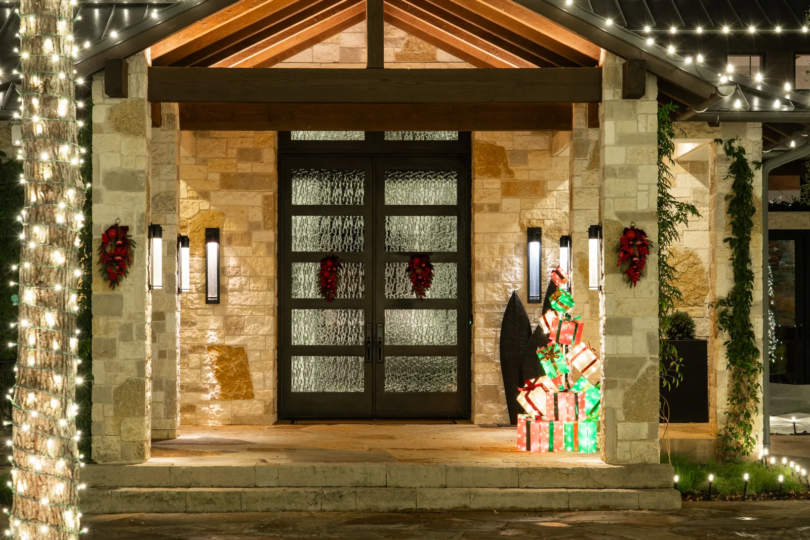 Stone home entryway at night with a tree trunk wrapped in warm white lights, fresh wreaths on the glass front doors, and illuminated wrapped gift-box light displays beside the porch.