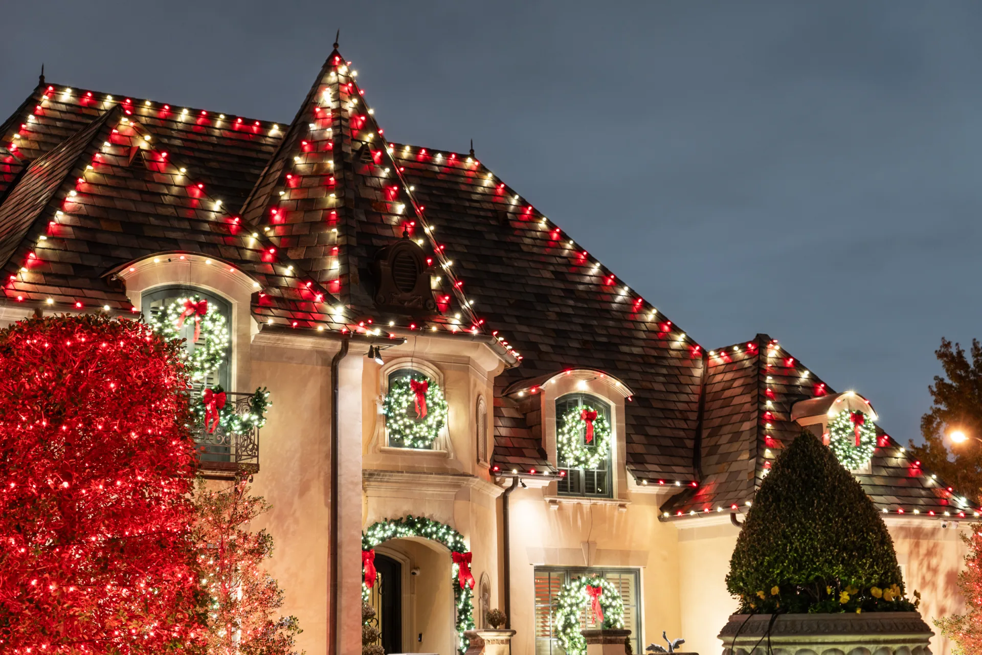 The Perfect Light Christmas lighting on a Texas storybook home with red and white roofline lights, window wreaths, and ivy-covered walls