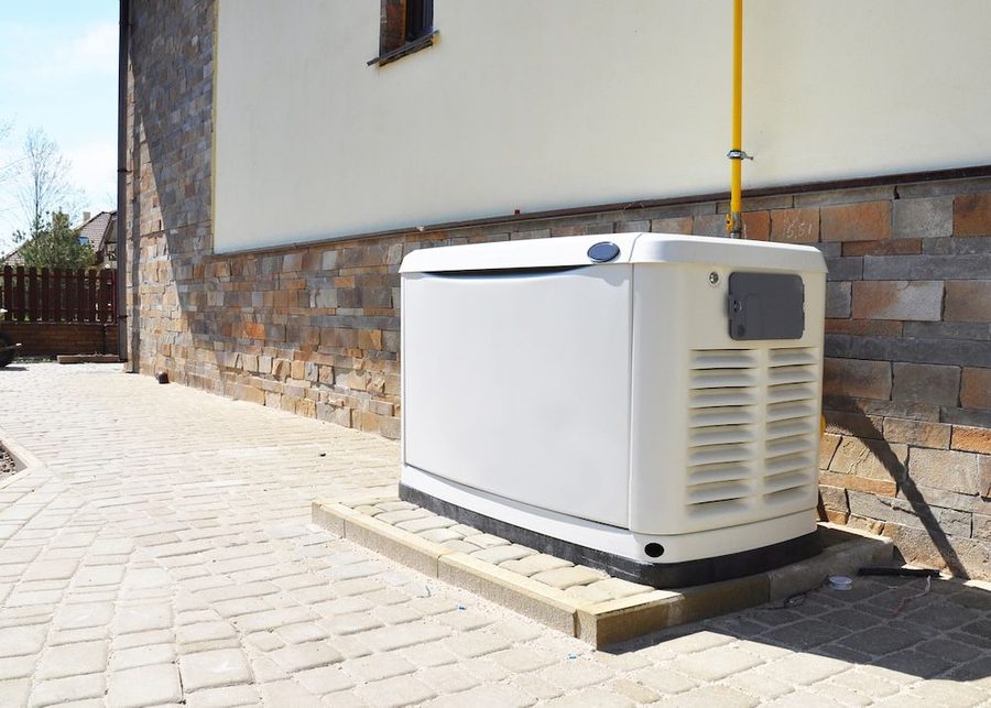 Outdoor HVAC condenser unit on a concrete pad beside a stone-wall house, along a brick-paved path.