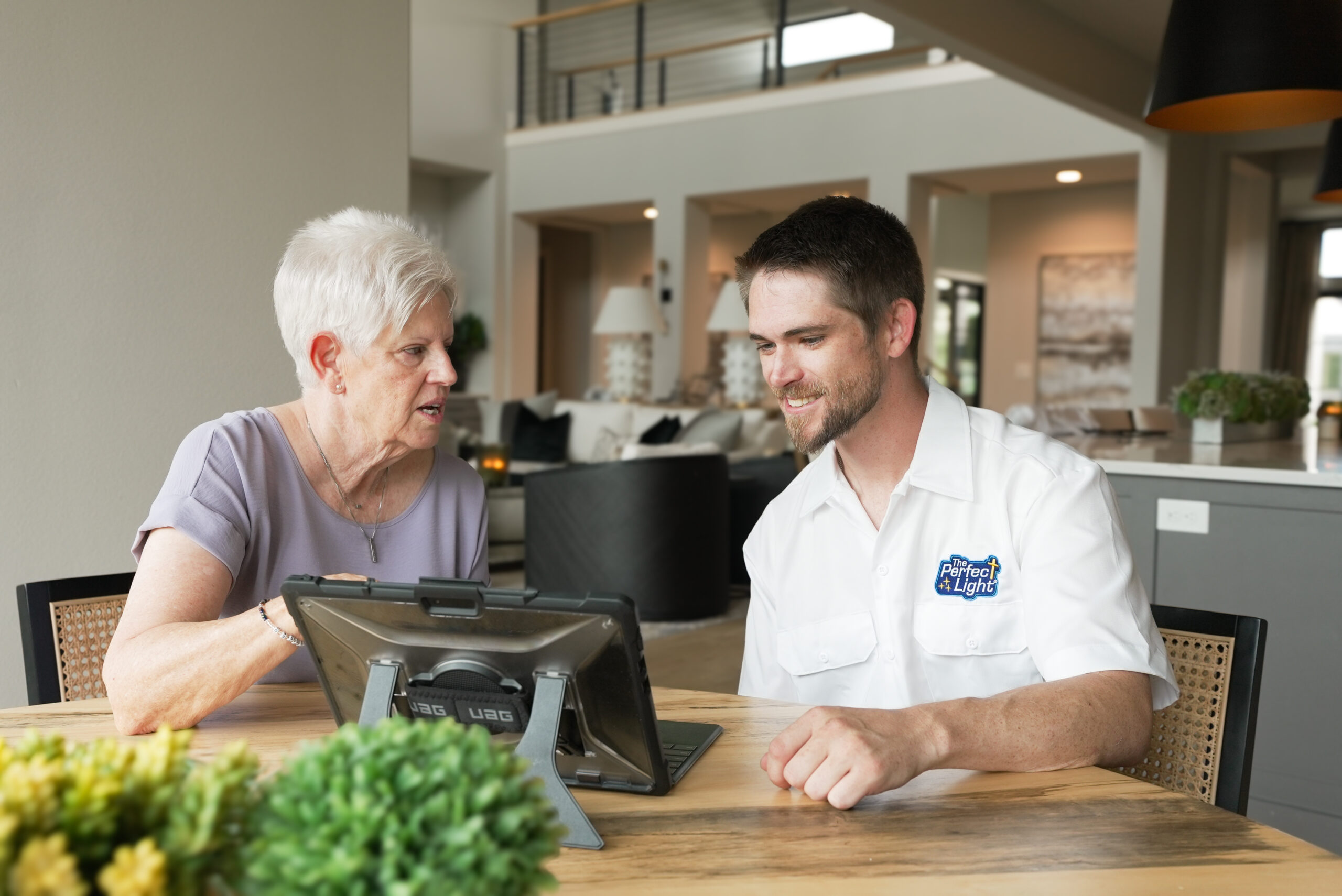 Older woman and man sit side by side at a wooden table in a bright showroom, looking at a tablet together.