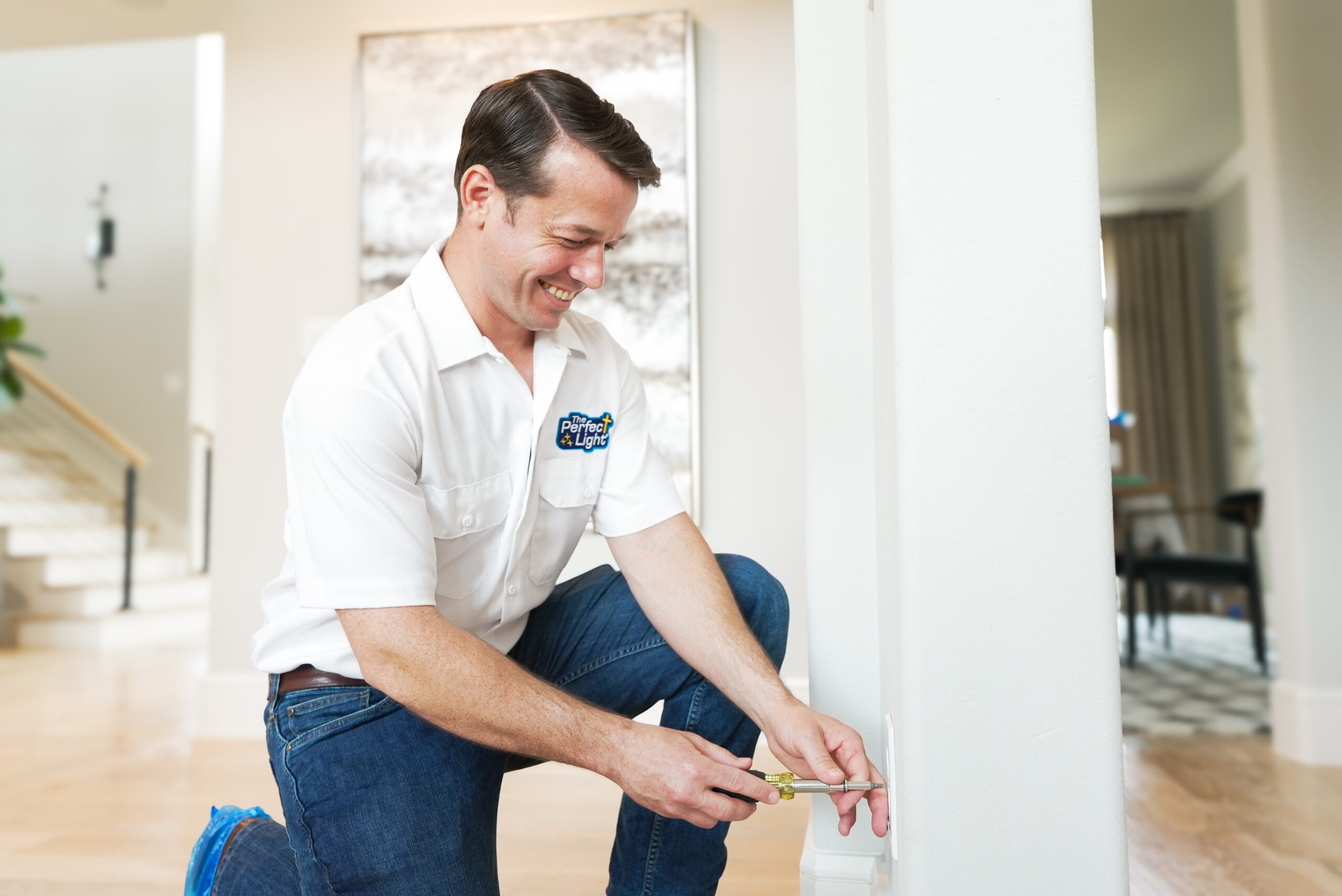 Man kneeling in a bright living area, installing or adjusting a wall electrical outlet with a smile on his face.